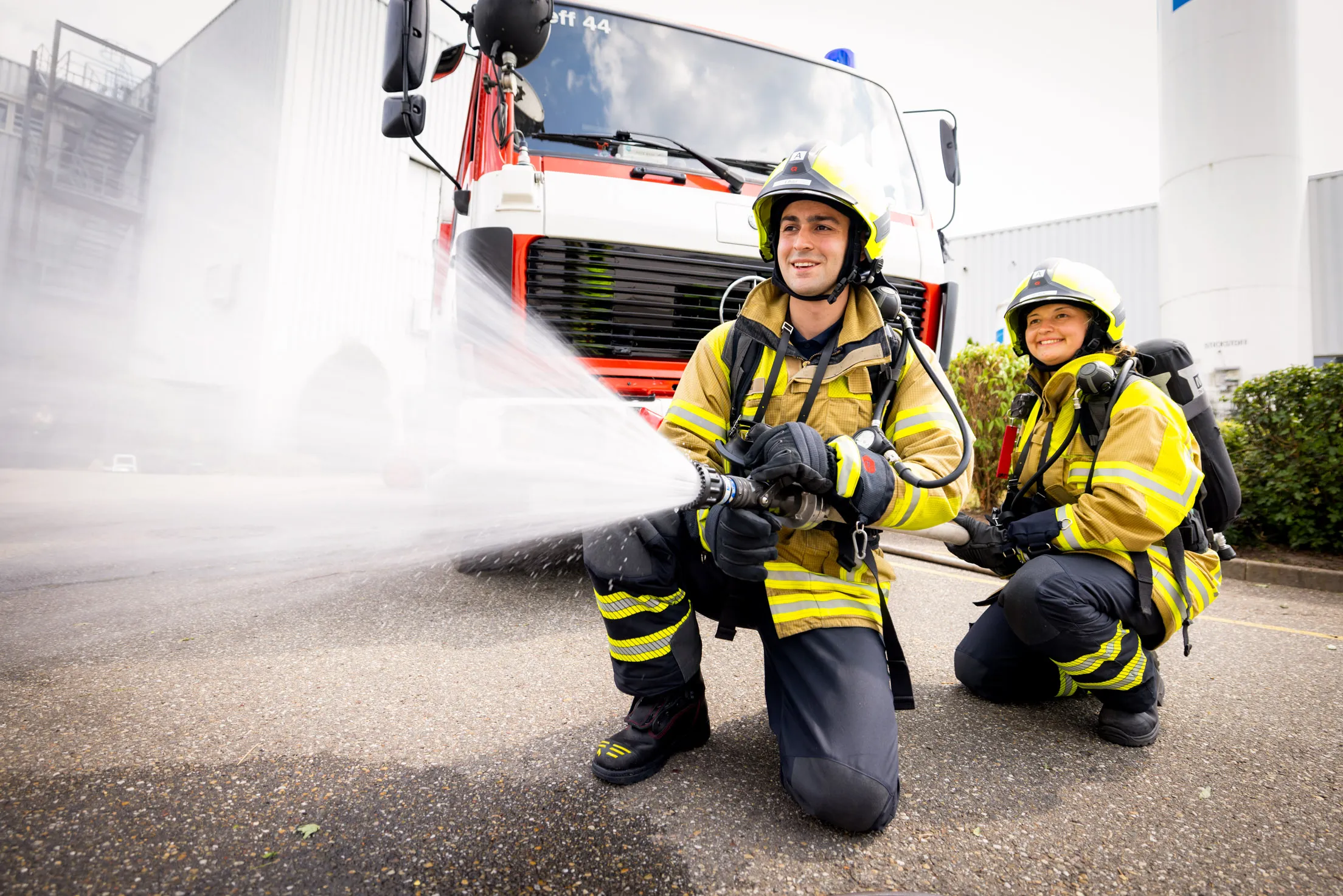 Zwei Personen der Feuerwehr in voller Montur mit gelbem Helm und Jacke, schwarzer Hose. Sie knien auf dem Boden, links ein Mann mit einem Feuerwehrschlauch der Wasser spritzt und daneben rechts eine Frau, die den Schlauch hält. Im Hintergrund ist unscharf ein Feuerwehrwagen zu sehen.