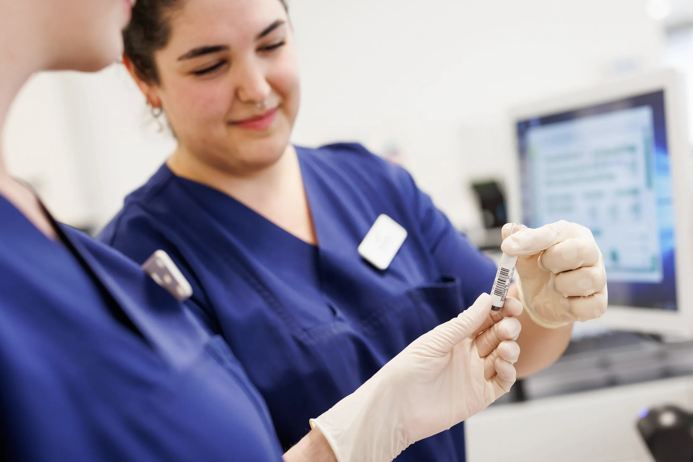 Zwei Mitarbeiterin in blauem Kasack mit weißen Handschuhen stehen eng zusammen, halten eine Blutprobe und sind in einem Labor zu sehen. Nur das Blutröhrchen ist scharf, alles andere ist unscharf. Die eine Dame ganz links ist nur im Anschnitt mit Oberkörper, Kinn und ihrer linken Hand sichtbar. Mittig im Bild steht die andere Frau mit Gesicht, schwarzem Haar und Oberkörper zu erkennen. Rechts im Foto befindet sich ein weißer Monitor mit blauer Schrift auf dem Display.