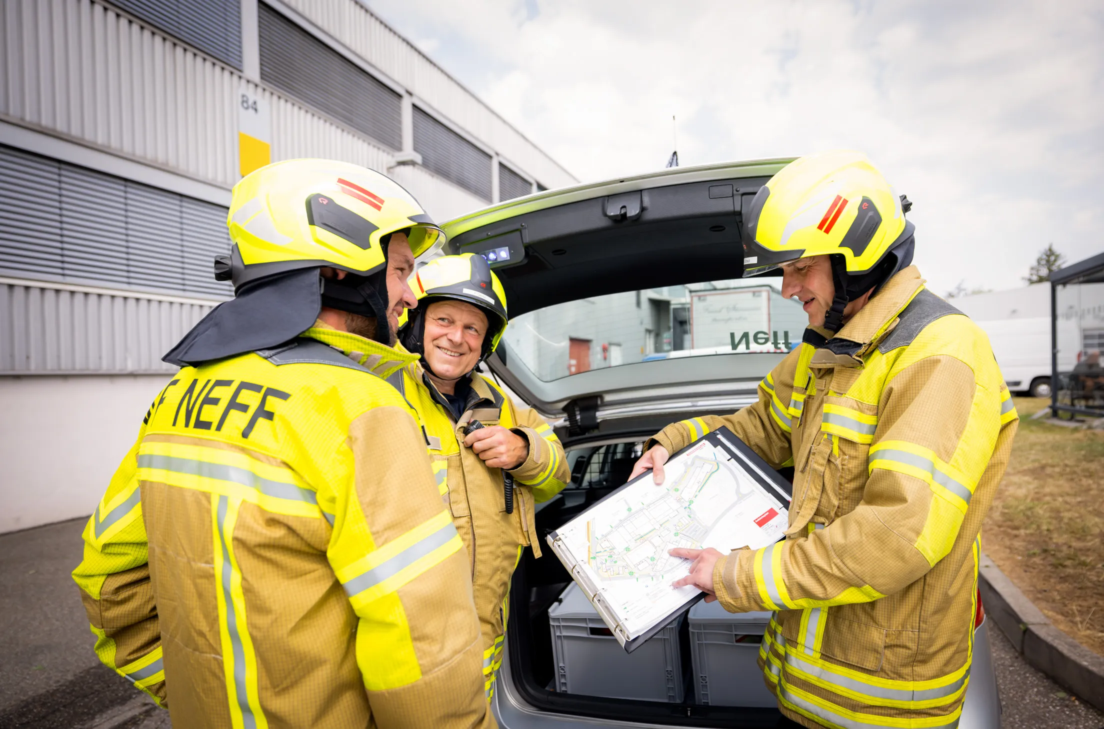 Drei Feuerwehrmänner in Montur stehen bei einem offenen Kofferraum eines Einsatzwagens und besprechen etwas. Sie tragen alle gelbe Feuerwehrjacken und gelbe Helme mit Visier. Der Feuerwehrmann rechts im Foto hält eine Mappe mit einem Lageplan, die beiden anderen Kollegen lachen sich an. Im Kofferraum sind zwei graue Kisten zu sehen.