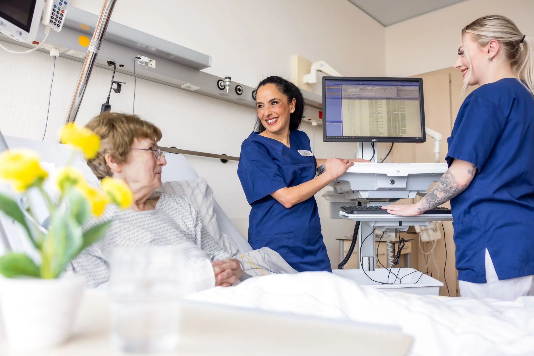 Blick in ein Patientenzimmer, links unscharf gelbe Blumen im Anschnitt und rechts hintern ein hellbrauner Schrank. Drei Personen sind zu sehen: eine Patientin links im Bild sitzt mit einem weißen Krankenhausnachthemd in einem Krankenbett, Kopflehne erhöht. Sie trägt aschblondes, kurzes Haar und eine Brille. In der Mitte steht eine Pflegerin mit schwarzem, langen Haar in einem Zopf an einem Visitenwagen und dreht sich zu der Patientin. Rechts am Bildrand eine blonde Krankenschwester mit Pferdeschwanz, von der Seite an dem Monitor stehend. Sie lächelt auch zu der Patientin. Beide Mitarbeiterinnen tragen weiße Hose und einen blauen Kasack.