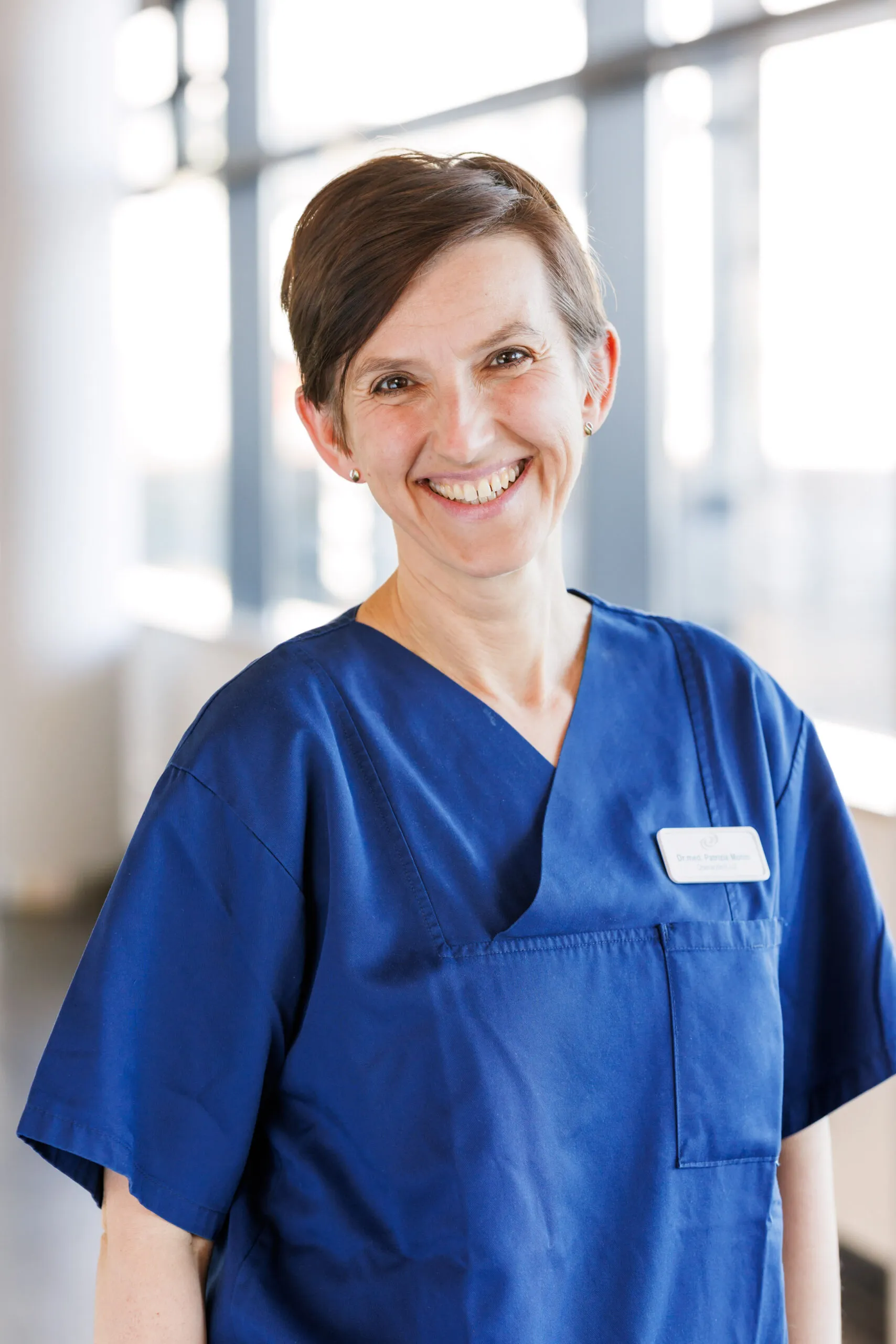 Hochformatiges Portrait einer Ärztin mittleren Alters, mit braunen, mittelkurzen Haaren und sie lacht in die Kamera. Sie trägt einen blauen Kasack und ist mit dem Oberkörper abgelichtet. Im Hintergrund ist unscharf ein heller Klinikgang mit Säulen und Fenster zu erkennen.