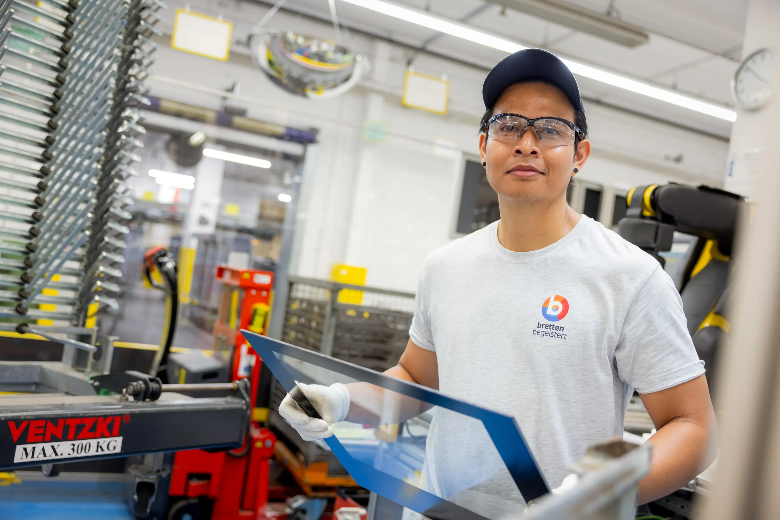 Ein asiatischer Mitarbeiter steht rechts im Bild mit hellgrauen Werksshirt, weißen Sicherheitshandschuhen und einer Sicherheitsbrille. Ausserdem trägt er eine dunkle Kappe und links und rechts dunkle, kleine, runde Ohrringe. Er hält eine Ofentür in der Hand und unscharf im Hintergrund ist die helle Werkshalle, Aufbewahrungsregale und ein roter Hubwagen zu sehen.