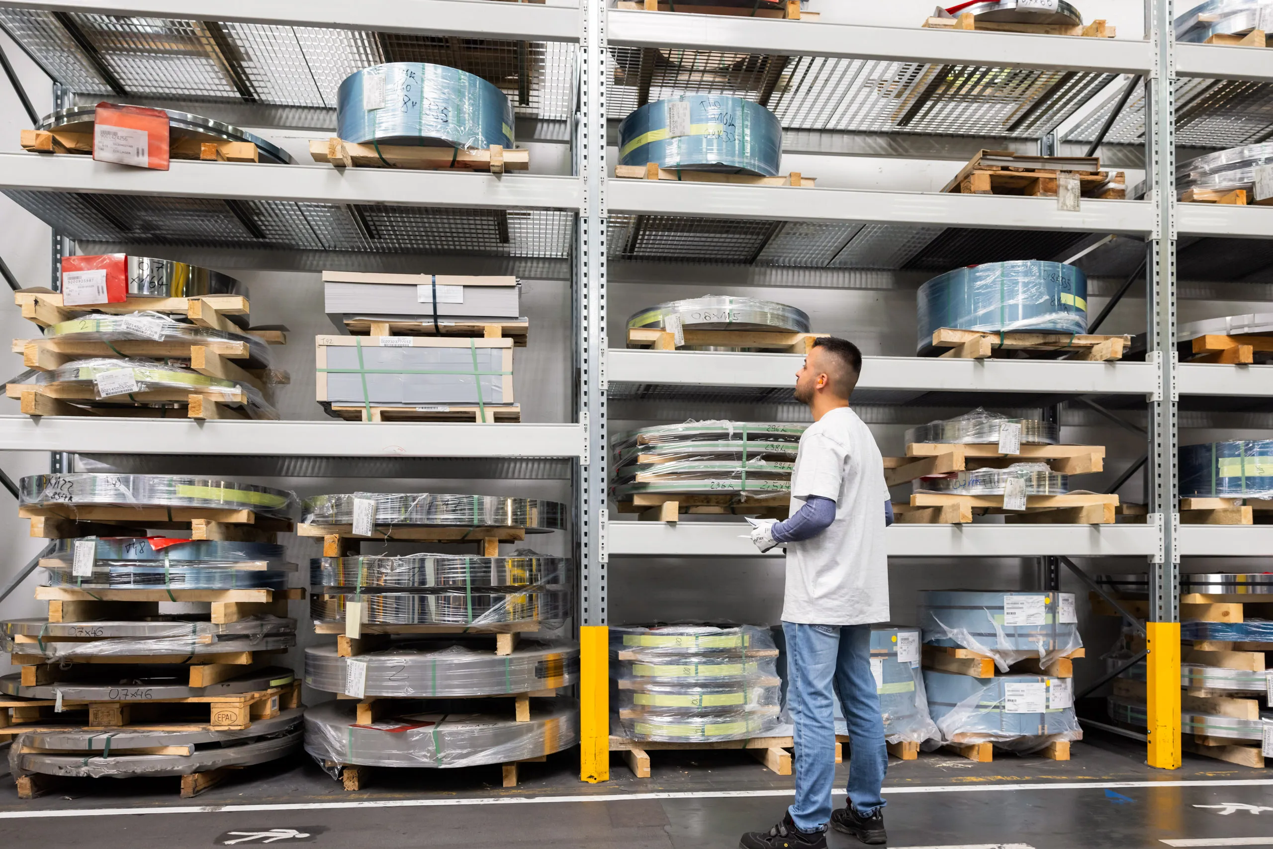 Ein Mann ist mit ganzem Körper, blauer Jeans, grauem T-Shirt, Sicherheitshandschuhen und Armschoner in der Logistik zu sehen. Er steht leicht nach links gedreht, auf der rechten Seite des Fotos und schaut auf ein großes, silbernes Regal mit abgepackten Zubehör, zusammengerollten Blechen in verschiedenen Größen. Die Ware ist auf verschiedene Holzpaletten gepackt.