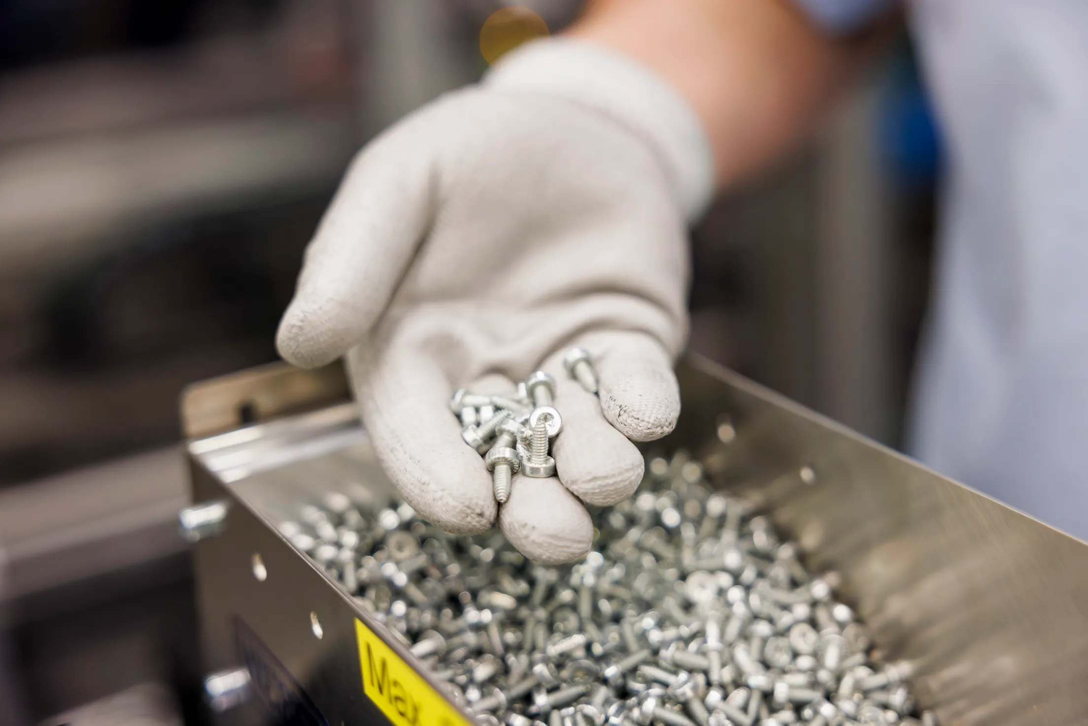 Ein Detailbild von einer Hand mit weißen Handschuhen, die viele kleine Schrauben hält. Unter der Hand ist eine silberne Kiste mit vielen weiteren Schrauben zu sehen.