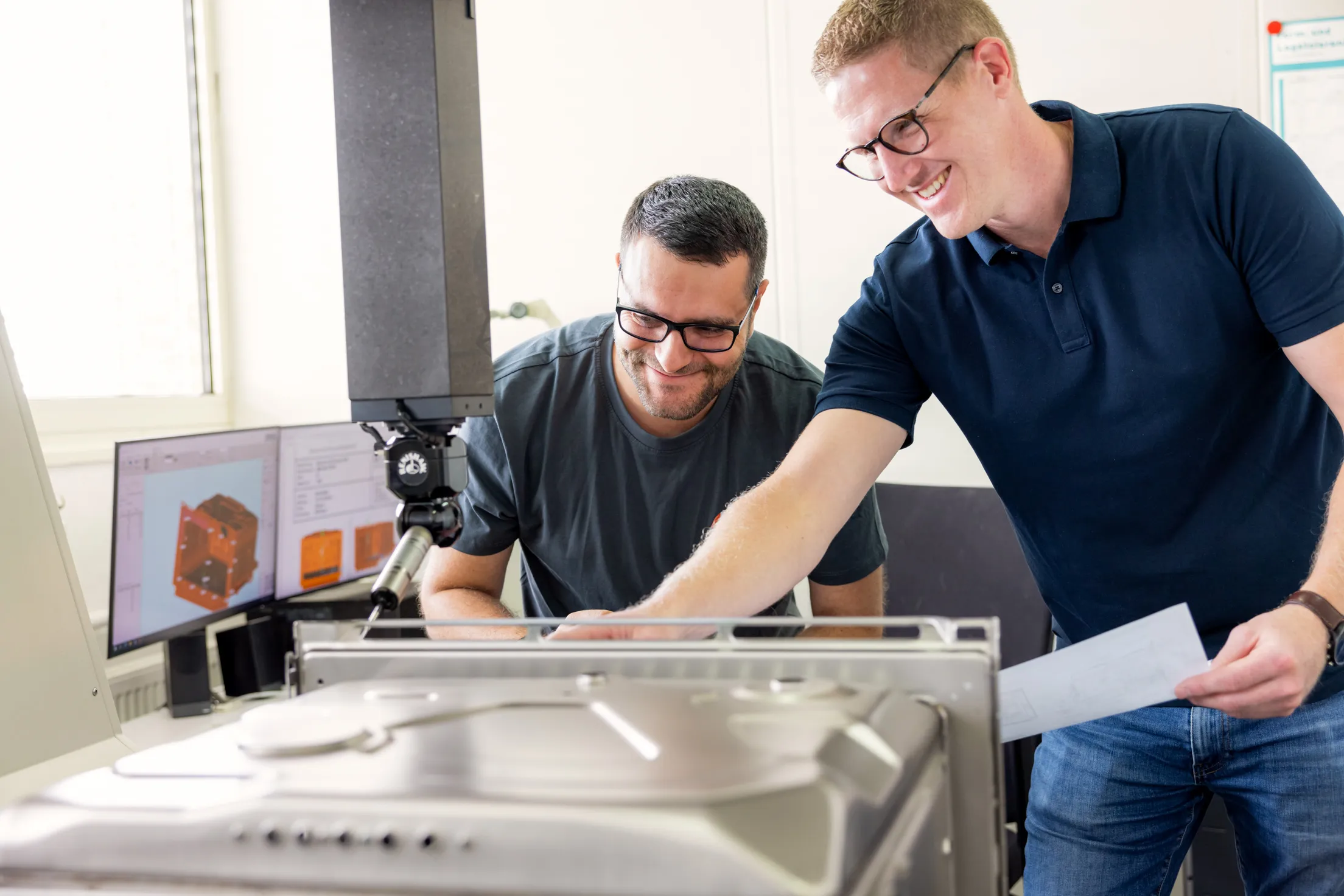 Zwei Männer aus der Entwicklung schauen auf ein silbernes Backofen Model, welches im Anschnitt mittig im Bild sich befindet. Ein Roboterarm bearbeitet den Ofen. Beide Männer tragen dunkle Brillen, rechts der Mann ein blaues Polohemd, Jeans und er hat blonde, kurze Haare. Der Mann links im Bild hat dunkle, kurze Haar, ein Vollbart und ein graues T-Shirt an. Links im Bild sind zwei Monitore mit orangen 3D Modellen in der Unschärfe zu sehen.