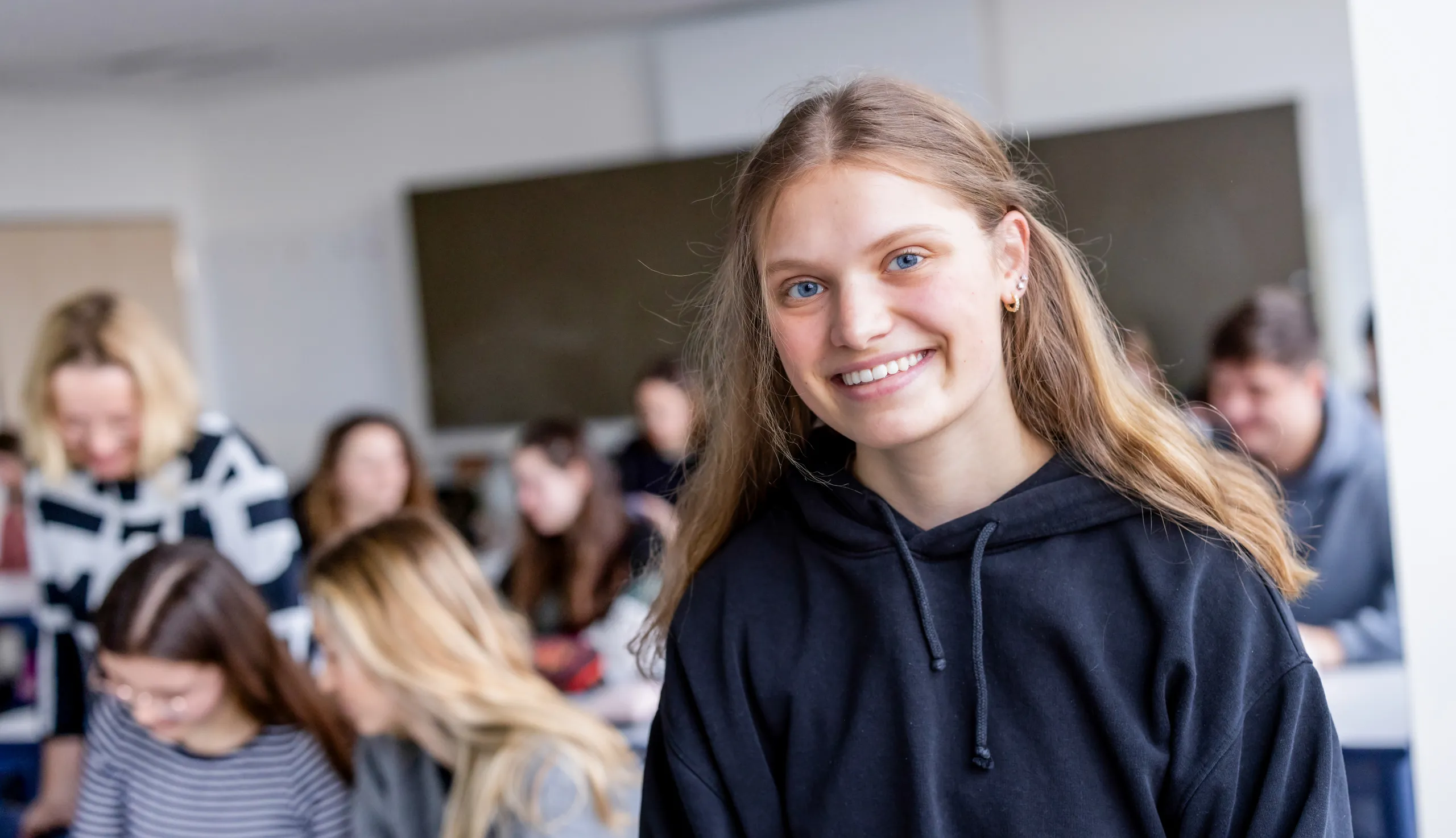 Eine Schwesternschülerin ist mit einem dunkelblauen Kapuzenpulli, langen dunkelblonden Haar, sehr blauen Augen und mehreren Ohrringen zu sehen. Sie sitzt auf einem Tisch und lächelt freundlich in die Kamera. Im Hintergrund unscharf ist das Klassenzimmer zu sehen mit einer großen, grünen Tafel in der Mitte und mehreren Schüler / -innen und einer Lehrerin, die sich mit dem Unterrichtsstoff beschäftigen.
