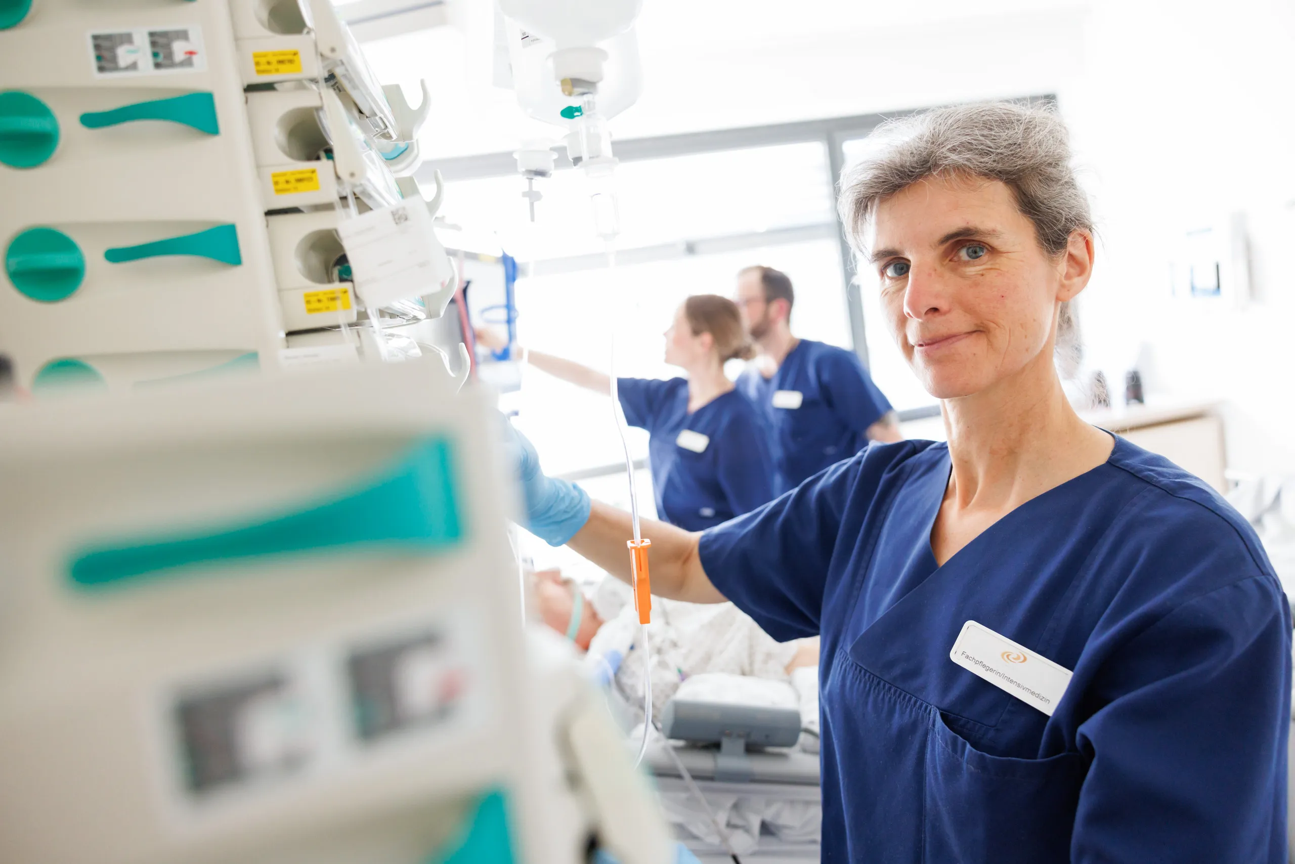 Drei Pflegekräfte mit dunkelblauen Kasack sind auf einer Intensivstation zu sehen. Links im Bild sind unscharf die Überwachungsgeräte in einem Zimmer der Intensivstation abgebildet mit Infusionen und Schläuchen. Rechts im Bild steht eine Pflegerin leicht angeschnitten mit hellblauen Handschuhen, sie schaut relativ ernst in die Kamera. Im Hintergrund ist ganz unscharf ein Patient im Bett und zwei Pflegekräfte an Geräten zu sehen.
