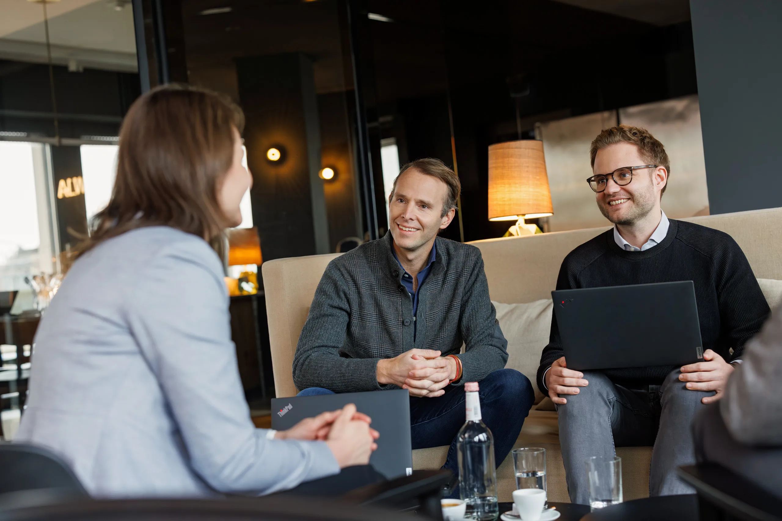 Eine dreier Gruppe von Personen, links sitzt eine Frau unscharf im Bild mit hellgrauer Jacke und auf einer hellen Couch sitzen zwei Männer. Der linke Mann trägt eine graukarierte Strickjacke und die rechte Person einen dunkelblauen Pullover und Brille, er hat einen Computer auf den Oberschenkeln. Die Menschen reden und lachen zusammen, im Hintergrund unscharf ein dunkler Raum mit einer Tischlampe und links im Bild ein Konferenzraum zu sehen