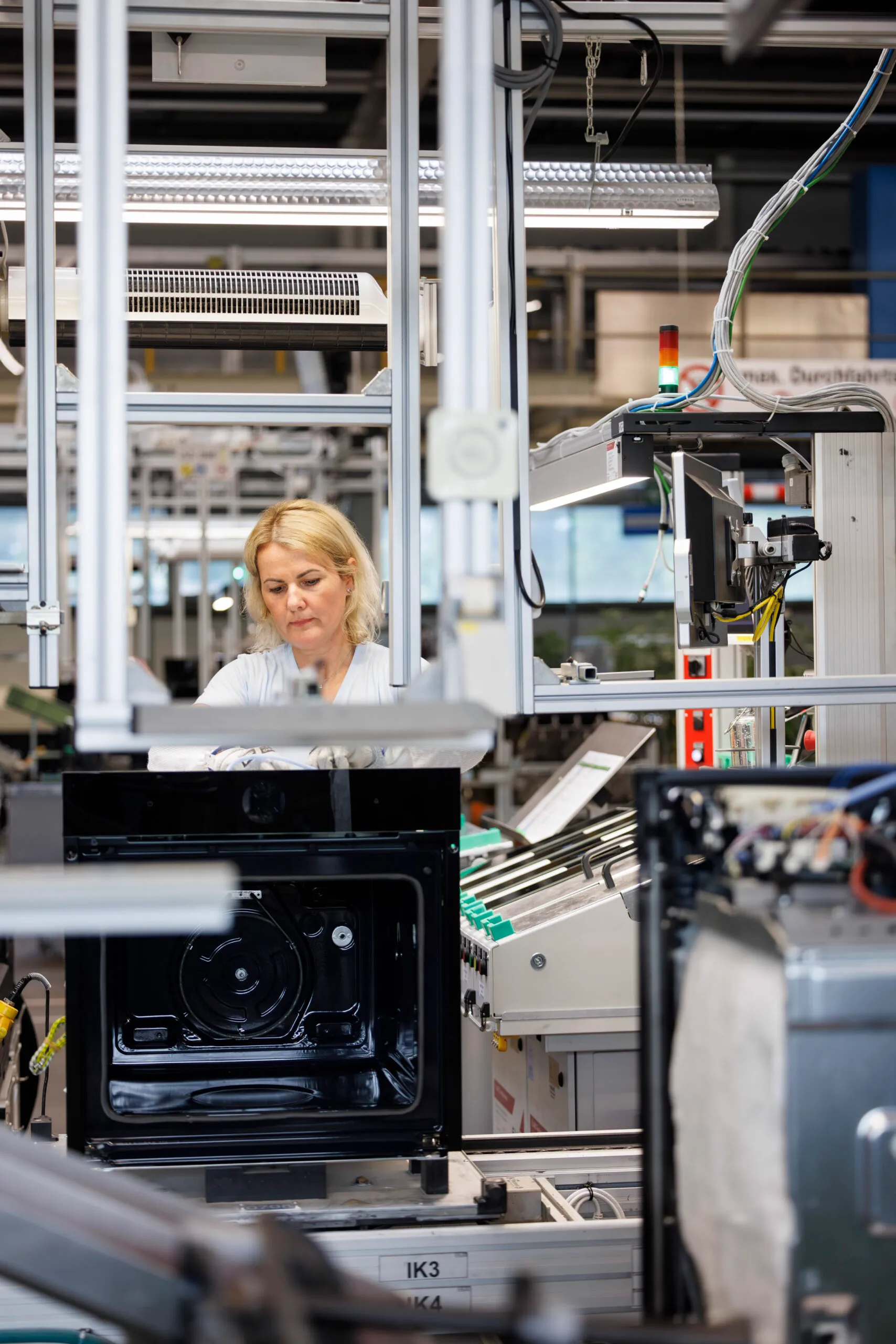 Eine Mitarbeiterin mit hellblauen Shirt, hat blonde kinnlange Harre und steht am Fließband. Sie arbeitet an einem Backofen.