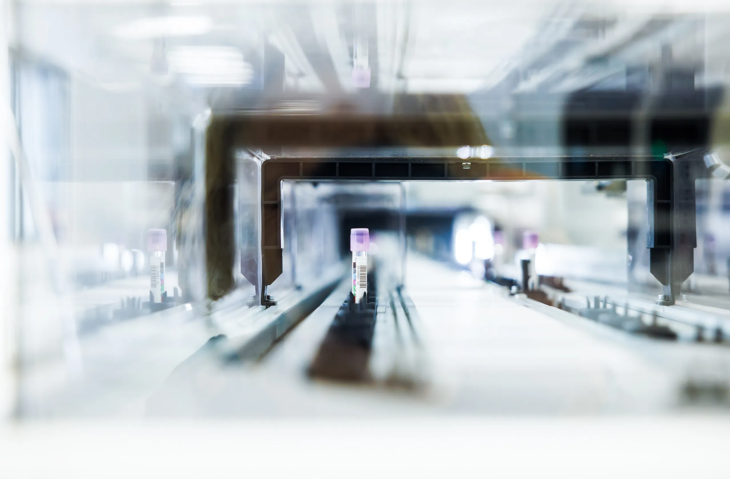 Ein Blutröhrchen mit lila Deckel und Barcode läuft durch eine Maschine. Das Röhrchen ist scharf, alles andere von der Maschine ist unscharf und in graublauen Tönen gehalten. Der Rest ist weiß von der Spiegelung.
