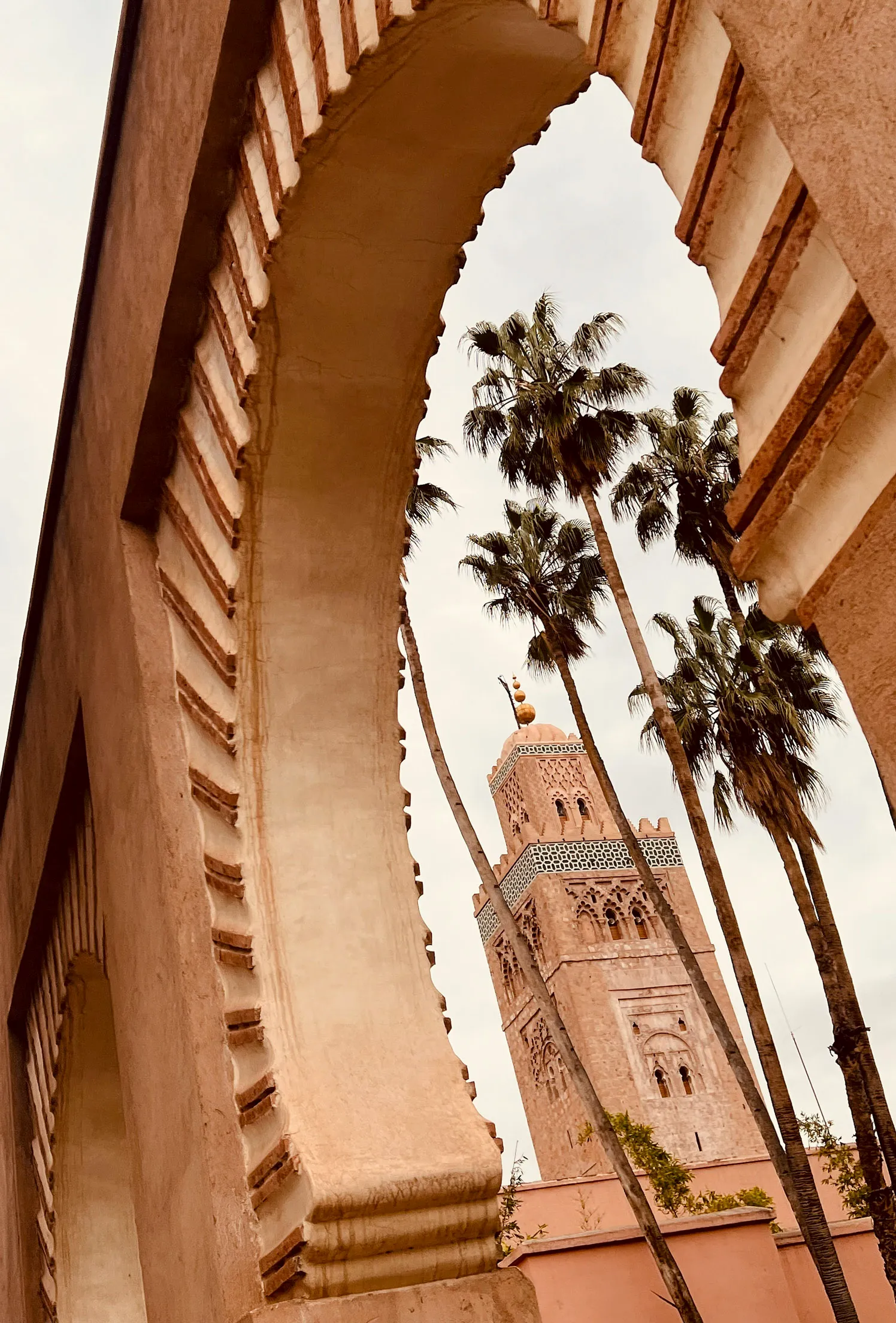 Durch einen Torbogen in Marrakech ist der Turm der Koutoubia Minaret zu sehen. Davor stehen ein paar Palmen. Das Foto ist von unten nach oben fotografiert.