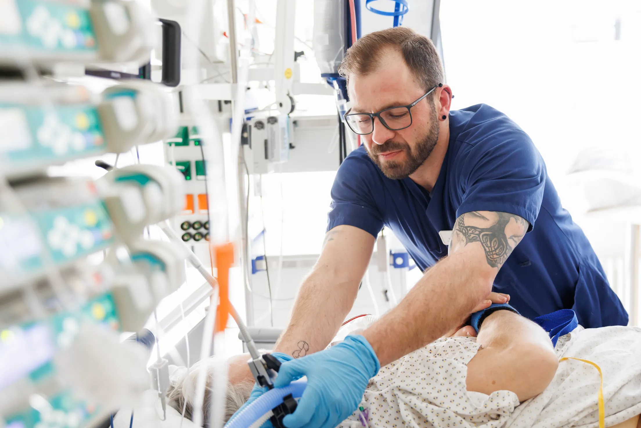 Auf der Intensivstation drückt gerade ein Pfleger mit dunkelblauen Kasack, hellblauen Handschuhen einen Knopf an Schläuchen die zu dem Patienten führen. Der Mitarbeiter ist an den Oberarmen tätowiert, trägt eine dunkle Brille und mehrere Ohrringe. Er beugt sich gerade über den Patienten, von dem nicht viel zu sehen ist, nur sein Kliniknachthemd und Unterarm ist zu erkennen. Links unscharf sind viele kleine Monitore mit türkisfarbenen Knöpfen zu sehen. 6./7./.8./ gehören zusammen