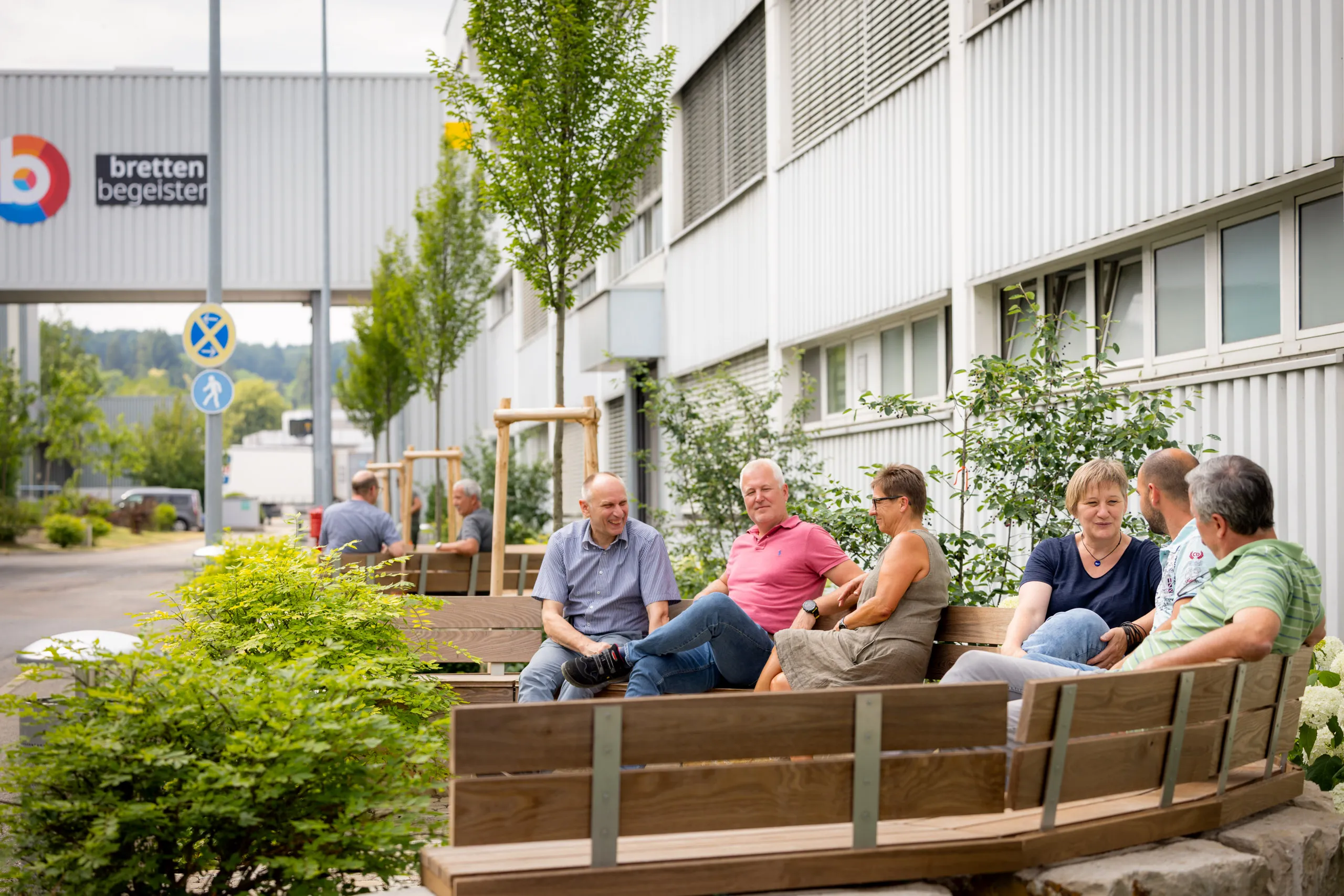 Eine Pausensituation draussen vor dem hellem Gebäude des Werks in Bretten. Das Logo ist unscharf auf einem großem Zwischengang links im Bild zu sehen. Das Gebäude erstreckt sich von links hinten nach rechts vorne und hat mehrere Fenster. Auf der rechten Seite sitzen mehrere Personen auf verschiedenen Holzbänken in 3-er Gruppen zusammen und unterhalten sich. Auf dem Werksgelände befinden sich viele Sträucher und Bäume in einem satten Grün.