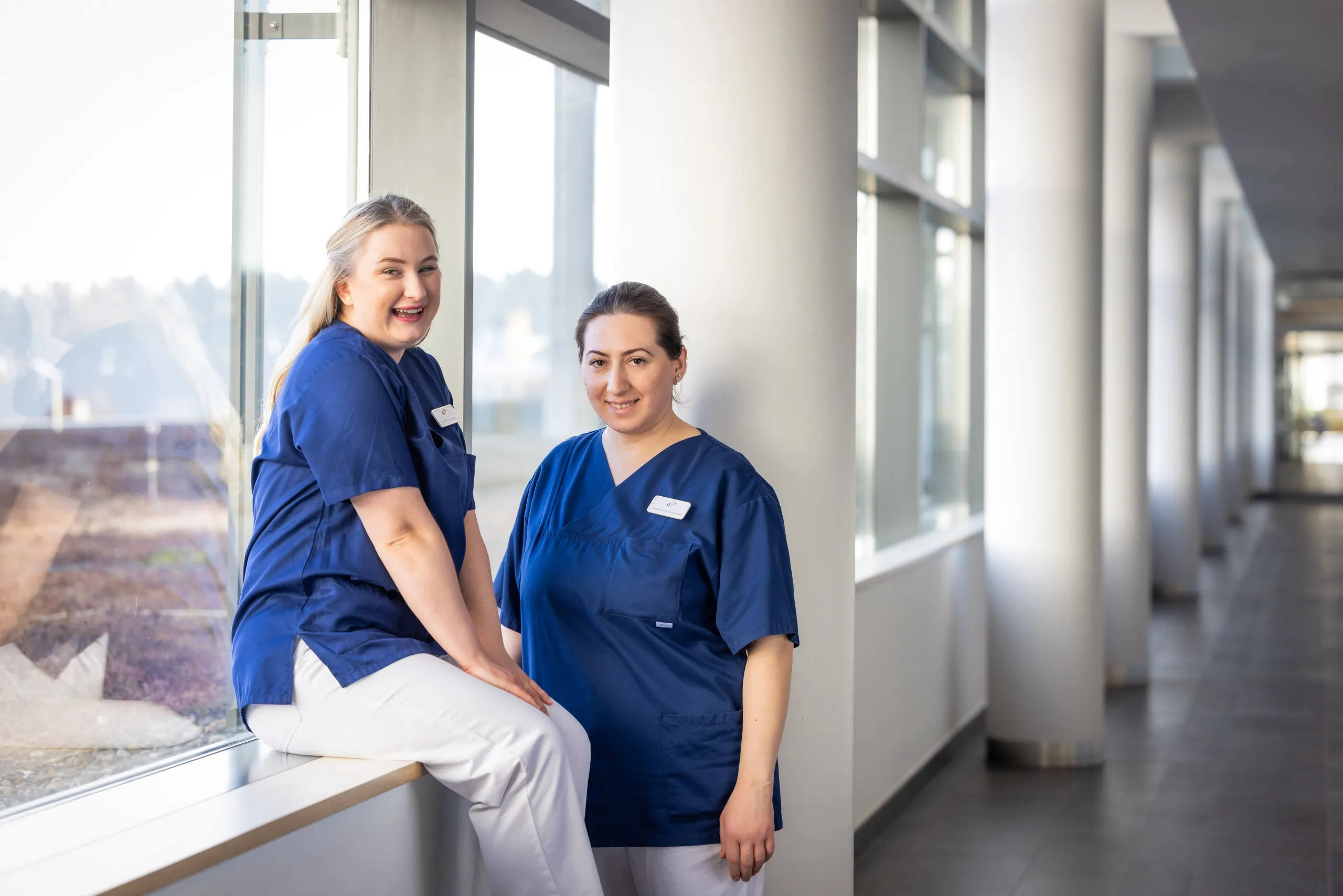 Ein Klinikgang mit Fenstern und Blick nach draussen, weißen Säulen in der Flucht und grauem Boden ist auf dem Foto zu sehen. Zwei Gesundheits- und Krankenpflegerinnen mit blauem Kasack und weißer Hose sind in der linken Fotohälfte. Die linke Dame sitzt lachend auf einer Fensterbank, sie hat blonde lange Haare, die Seiten zusammengebunden. Die Kollegin steht leicht lächelnd daneben und hat dunkle Haare zu einem Zopf gebunden. Beide Frauen schauen in die Kamera.