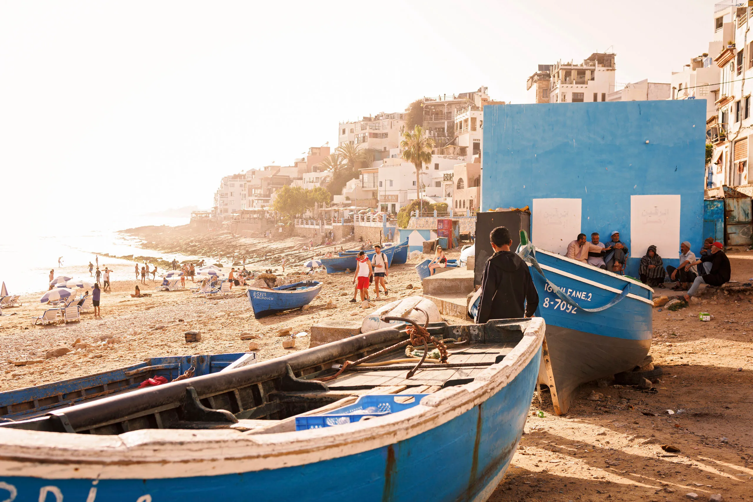 Blick auf den Hafen von Taghazout in Marokko an der Atlantikküste. Es sind blaue Fischerboote zu sehen und Menschen am Strand . Im Hintergrund ist die Stadt im Gegenlicht zu sehen.