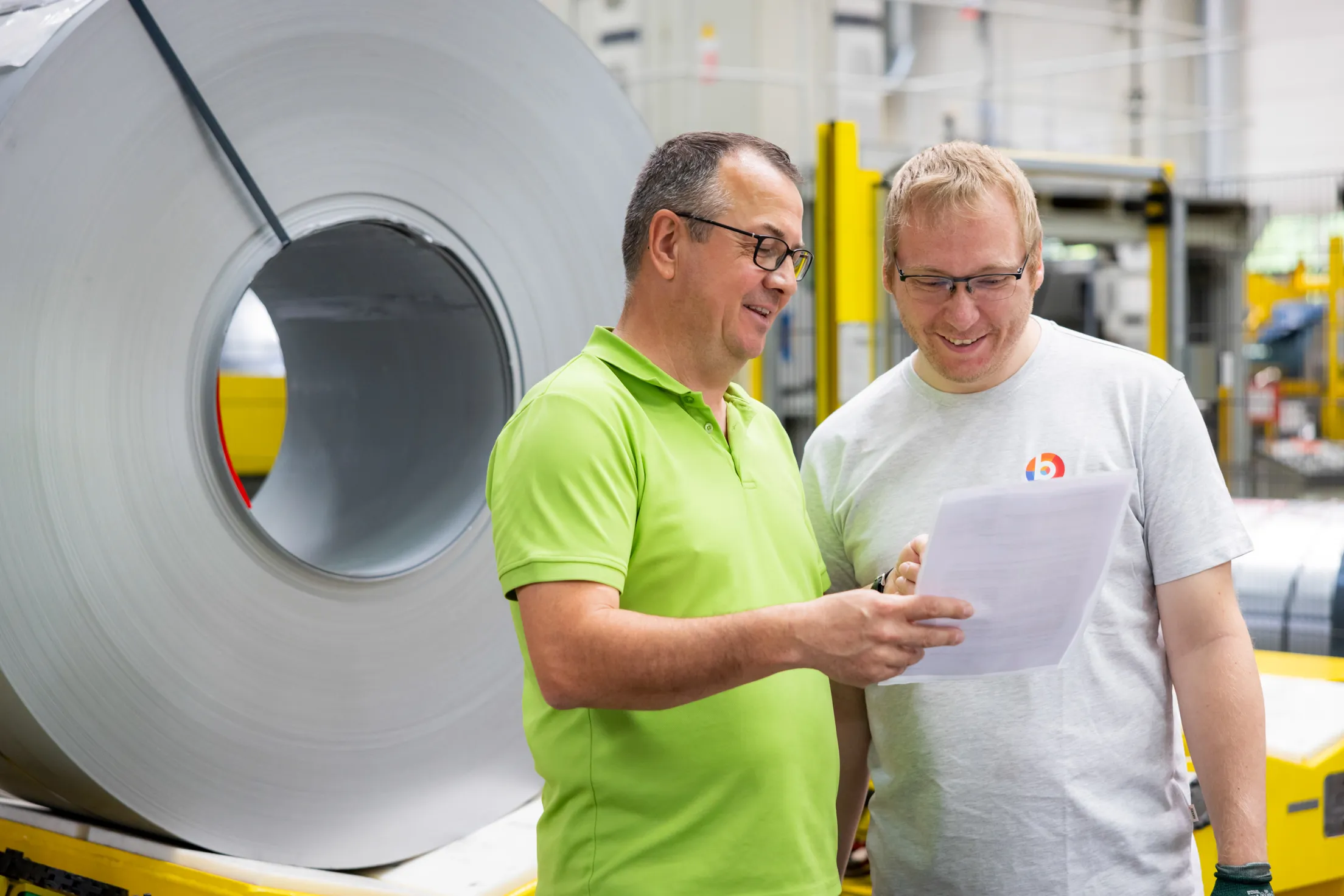 Zwei Männer mittleren Alters stehen in der Fertigung vor einer großen, silbernen Rolle und besprechen etwas. Beide Herren lachen dabei. Der Mann links mit grünem Polohemd, leicht ergrautem Haar und schwarzer Brille hält ein Blatt Papier in der Hand, der andere Mitarbeiter trägt ein graues BSH T-Shirt, hat blonde Haare und auch eine Brille.