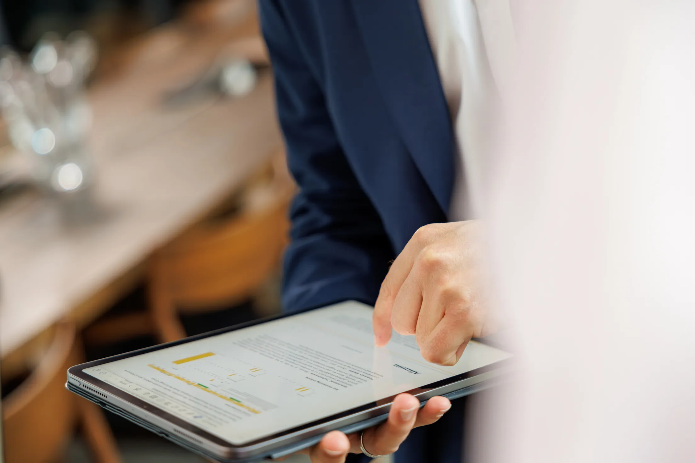 Zwei Hände halten ein silbernes Tablet und Finger scrollen einen Text. Die Person trägt ein weißes Hemd und ein dunkelblaues Sakko, aber im Anschnitt und Unschärfe zu erkennen. Ausserdem noch im Hintergrund ein Holztisch mit braunen Stühlen unscharf abgebildet.