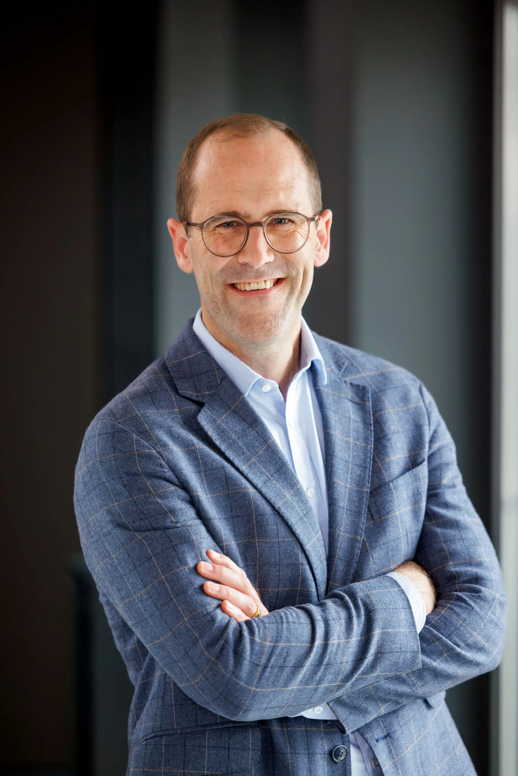 Portraits von der Geschäftsführung, ein Mann mittleren Alters, Brille und braunem kurzem Haar. Er trägt ein hellblaues Hemd, ein blau kariertes Sakko und lacht in die Kamera. Im Hintergrund dunkle, unscharfe Strukturen in Schwarz und Grautönen.