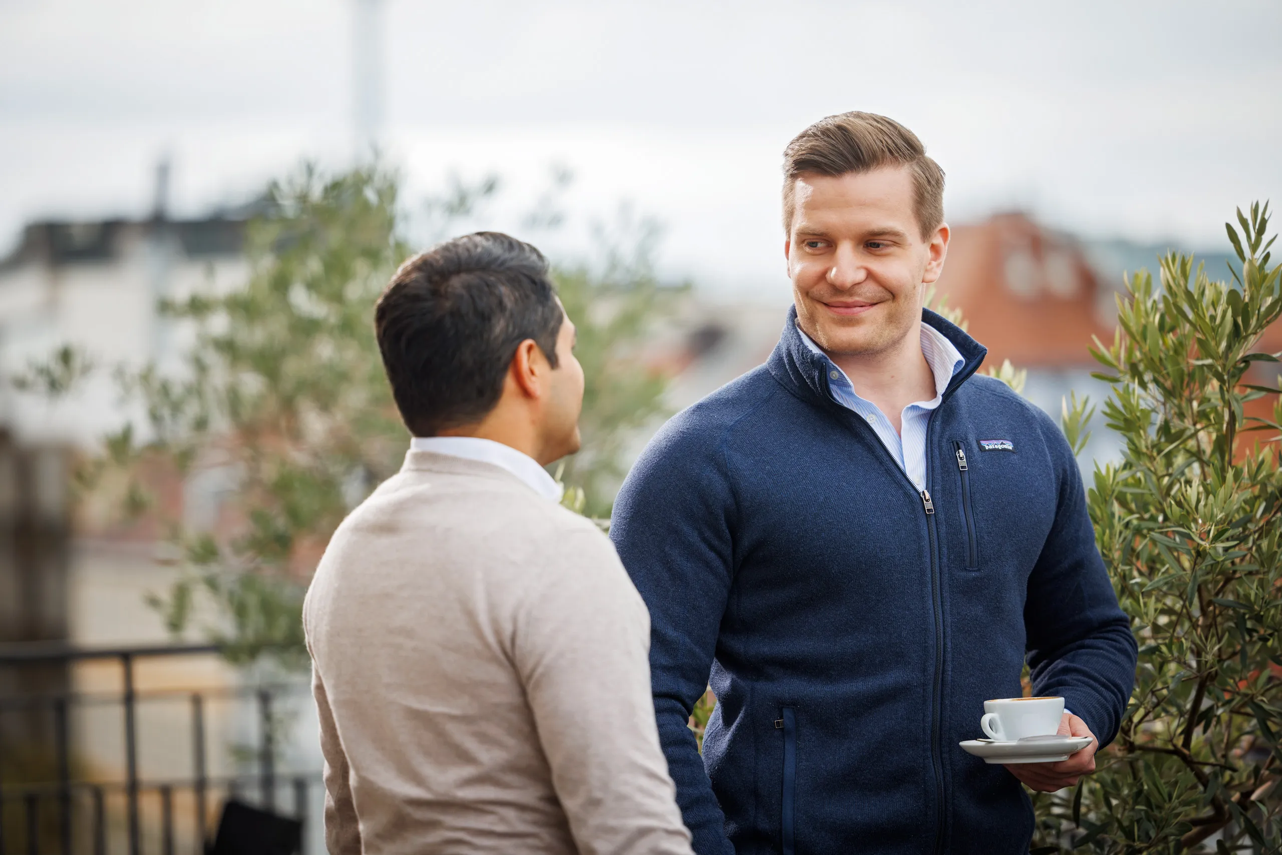 Zwei junge Männer stehen im Pullover auf einer Dachterrasse und unterhalten sich. Im Hintergrund Pflanzen und unscharf die Kulisse von München zu erkennen. Der rechte Mann trägt ein hellblaues Hemd mit einem dunkelblauen Pullover und hat eine weiße Kaffeetasse in der Hand. Er hat hellbraune kürzere Haare und lächelt seinen Kollegen an. Dieser trägt ein weißes Hemd, beigen Pullover und steht seitlich gedreht zur Kamera. Er hat kurze schwarze Haare und schaut seinen Kollegen an.