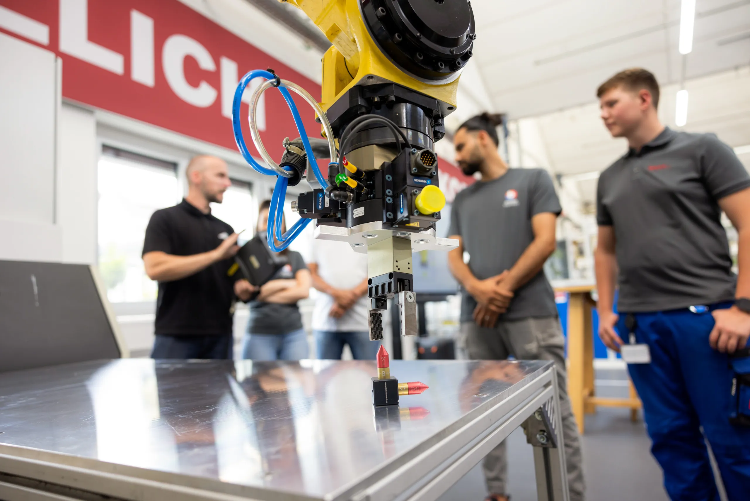 Im Vordergrund ist ein gelb, schwarzer Roboterarm mit einem Greifarm scharf abgebildet. Das Ganze über einem silbernen Metalltisch. Im Hintergrund ist in der Unschärfe eine Gruppe von fünf Personen in einer Werkstatt zu sehen. Ein Mann auf der linken Seite erklärt etwas, die anderen Personen sind Auszubildende in ihrer Arbeitskleidung. Links oben im Bild ist ein rotes großes Schild mit weißer Schrift zu sehen.