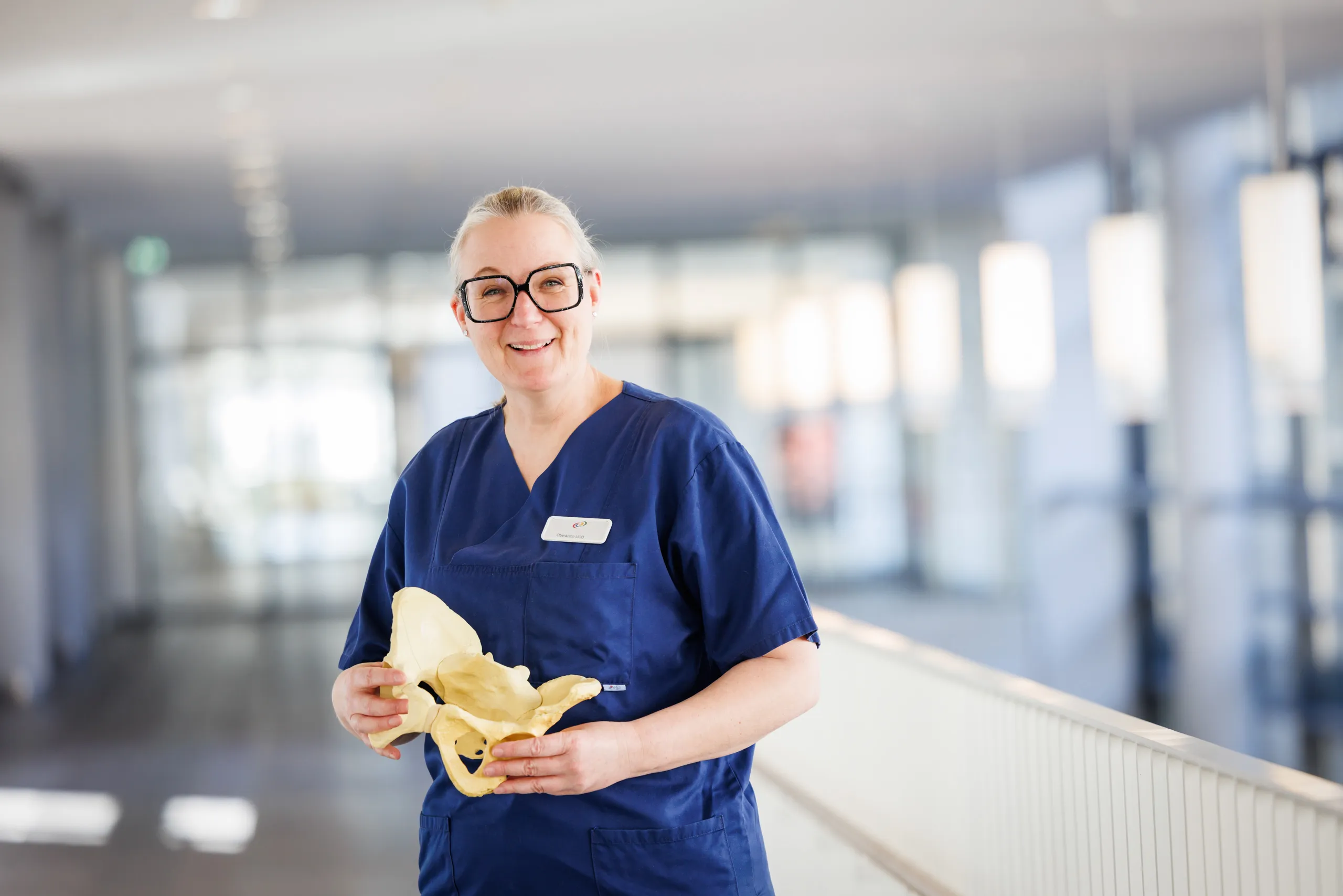 In dem Foto ist eine blonde Ärztin mittleren Alters zu sehen. Sie trägt ihre Haar streng nach hinten und hat eine auffällige, schwarze, quadratische Brille. In den Händen hält sie ein Hüftmodell und sie hat einen dunkelblauen Kasack an. Im Hintergrund total unscharf ein heller Klinikgang in Blautönen mit Säulen, vielen Lampen, Glastüren und grauem Boden. Rechts im Bild ein weißes Geländer.