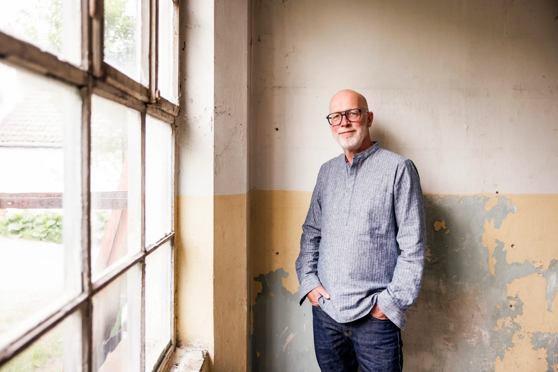 Mann mittleren Alter steht lässig an einer Wann in einem leeren Raum mit alter Fensterfront. Er hat keine Haare, trägt eine Brille und schaut leicht lächelnd in die Kamera
