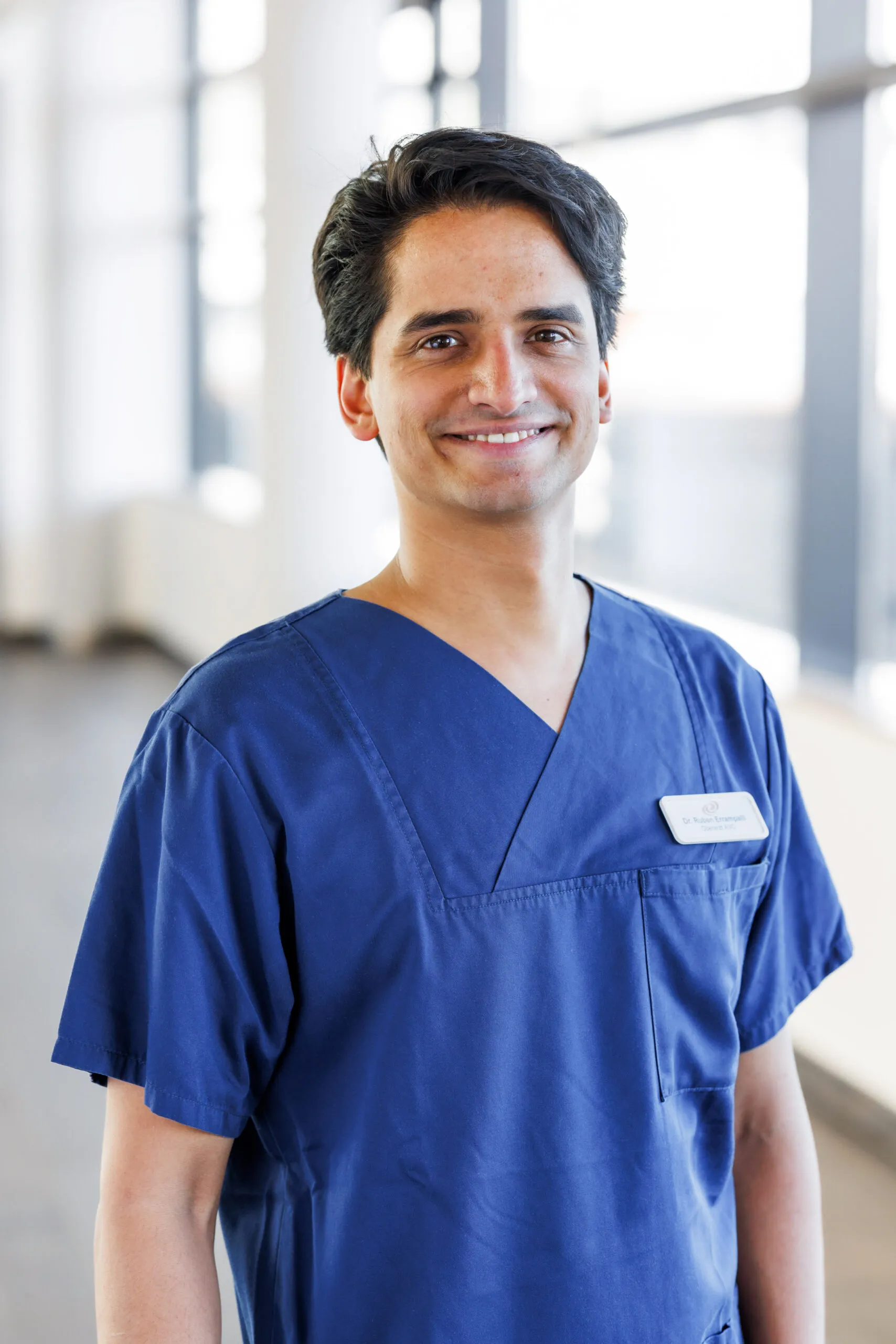 Hochformatiges Portrait eines jungen Arztes aus Asien. Er hat dunklere Haut, schwarze kürzere Haare und lächelt in die Kamera. Er trägt einen blauen Kasack und ist mit dem Oberkörper abgelichtet. Im Hintergrund ist unscharf ein heller Klinikgang mit Säulen und Fenster zu erkennen.