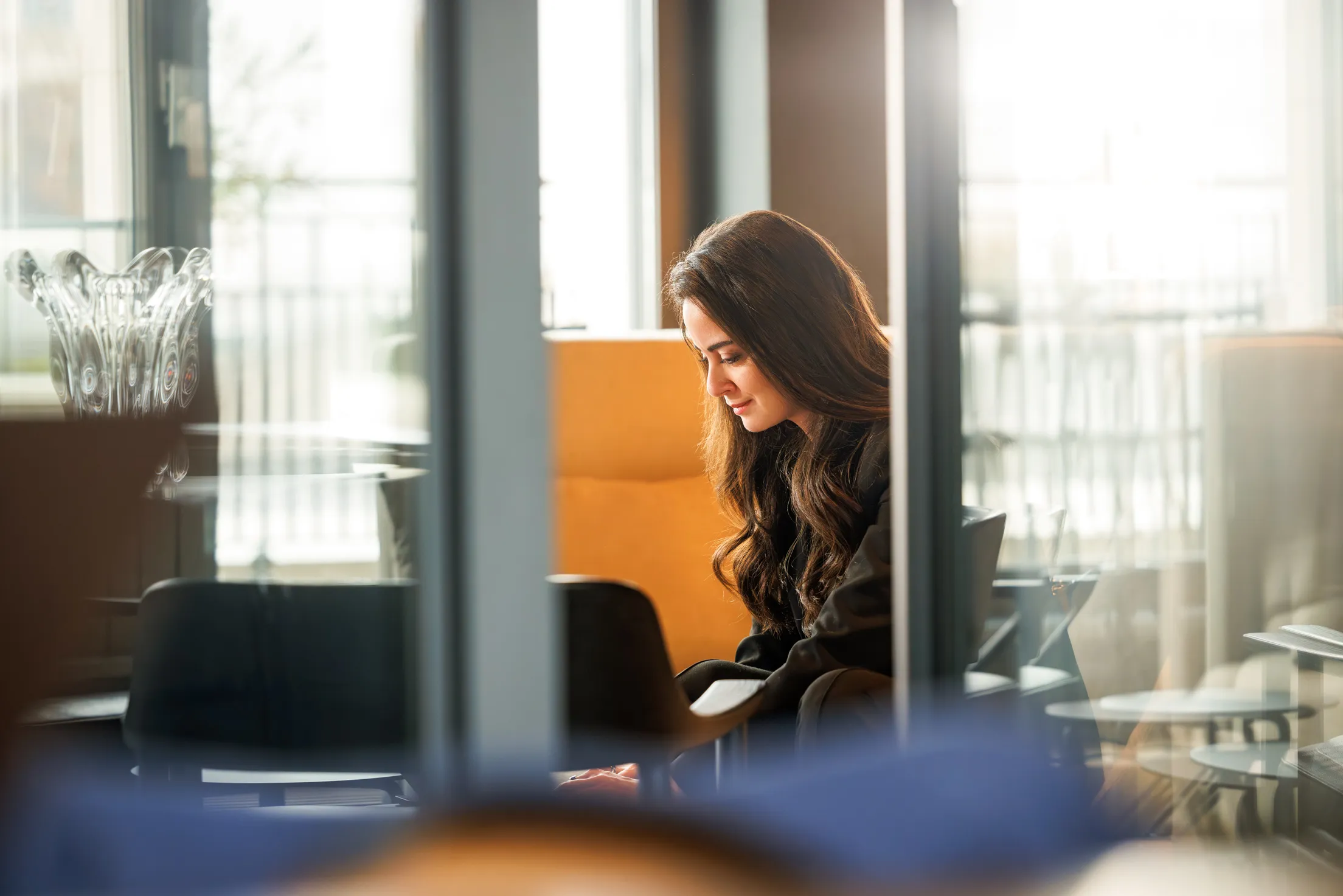 Durch eine Glastür sieht man eine Frau sitzend mit einem schwarzen Jacket und mit schwarzen langen, welligen Haaren. Sie liest etwas. Viel Spieglung in den Glasflächen, ein beiger Sessel und schwarze Stühle unscharf vor der Fensterfront zu sehen.